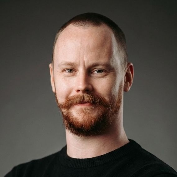 Portrait of a fair-skinned man with short, red hair and a full red beard and mustache, wearing a black sweater, set against a grey background.