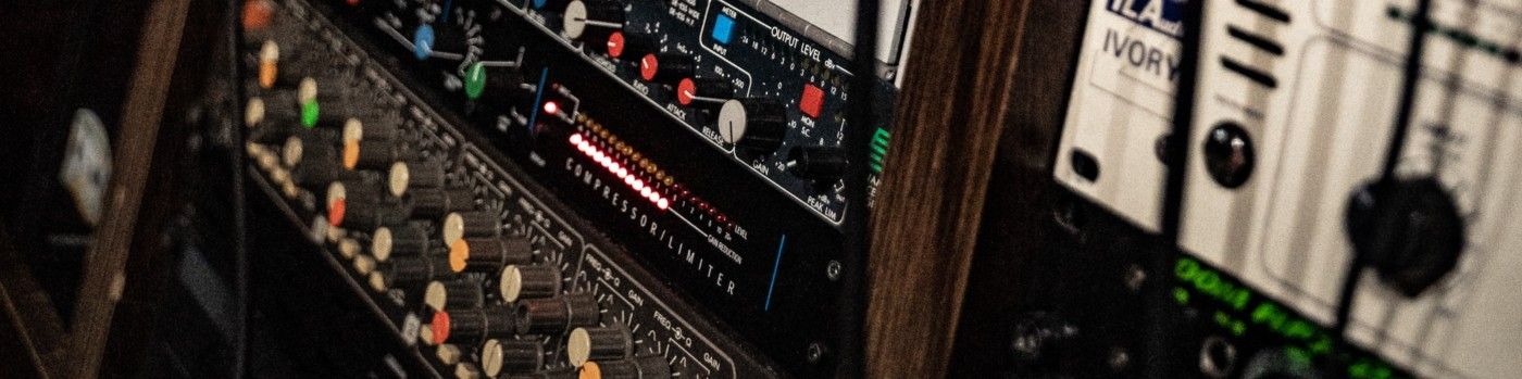 Close-up of professional audio equipment rack, featuring a compressor/limiter unit with lit gain reduction LEDs, alongside other audio processors with knobs, dials, and illuminated indicators, all mounted in a wooden rack.