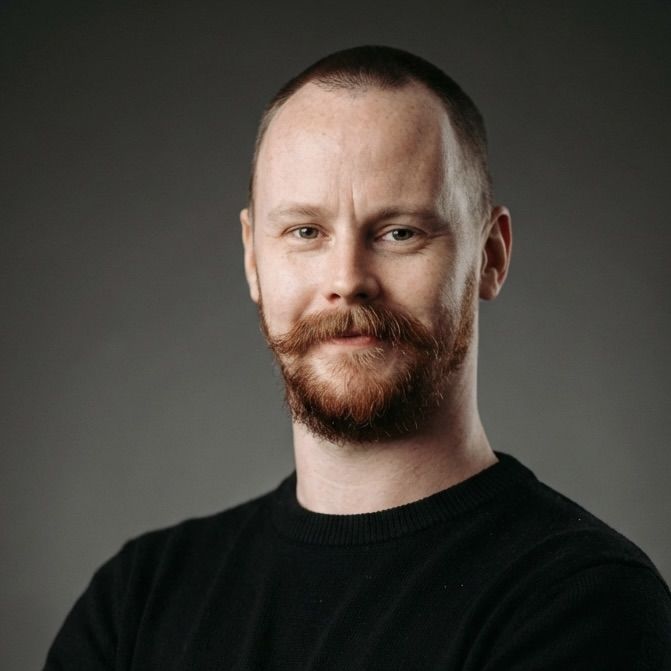 Portrait of a fair-skinned man with short, red hair and a full red beard and mustache, wearing a black sweater, set against a grey background.